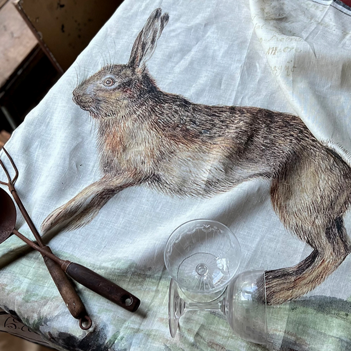 Barbara O'Donovan XL linen tea towel printed with a leaping brown hare, styled on a table with antique glassware
