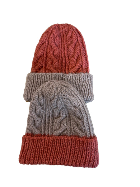 Two cable-knit beanie hats stacked on a patterned fabric surface. The top hat is coral red with a ribbed cuff, and the bottom hat is light taupe with a coral red ribbed cuff.