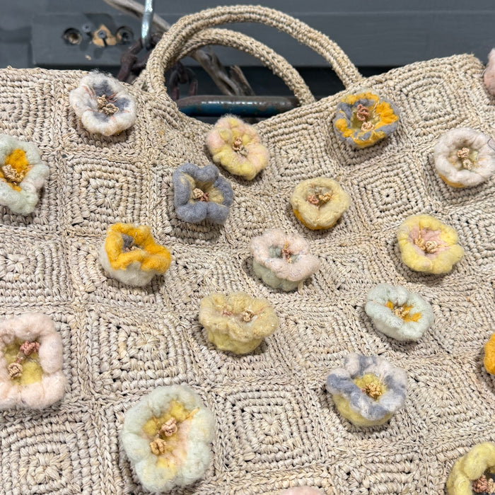 A woven straw tote bag with a geometric pattern, decorated with multiple textured, flower-like embellishments in pastel yellow, pink, blue, and white, resting on a rustic wooden surface.