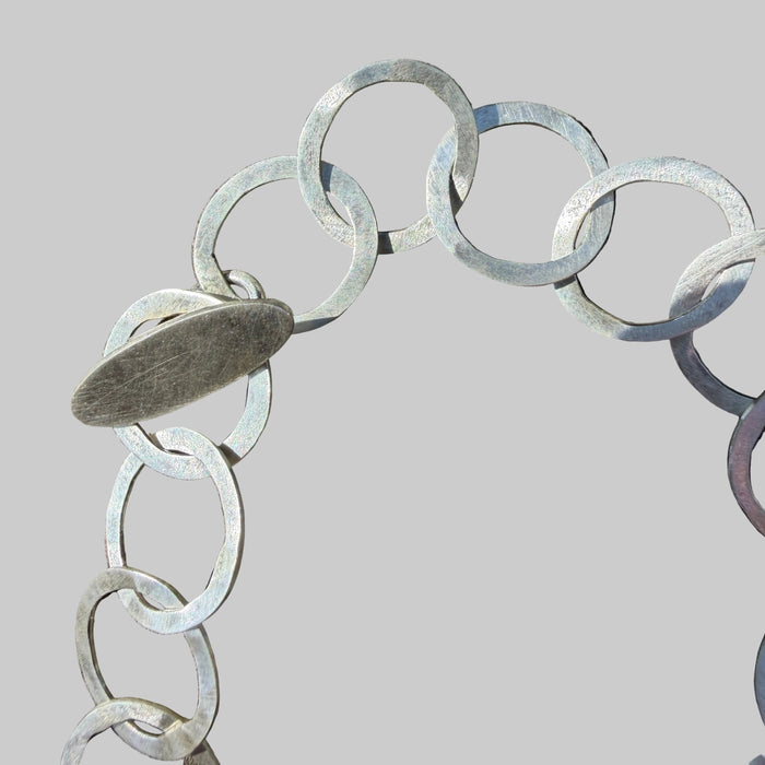 A close-up of a silver-toned chain bracelet with flat circular links and a single oval tag, photographed on a weathered wood surface with strong sunlight and shadows.