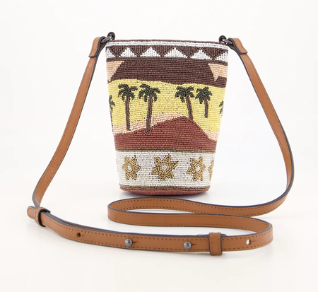 A hand-beaded bucket bag with a brown leather shoulder strap, featuring a Southwestern-style design with palm trees, hills, and geometric patterns in brown, yellow, white, and black.