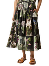 A model wears a black midi skirt featuring a botanical patchwork print with various flowers and leaves in green, pink, and white. The skirt has side pockets and is paired with brown slide sandals with colorful beaded straps, set against a plain light gray studio background.
