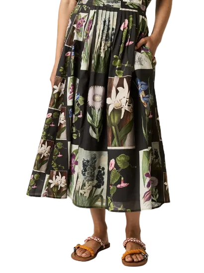 A model wears a black midi skirt featuring a botanical patchwork print with various flowers and leaves in green, pink, and white. The skirt has side pockets and is paired with brown slide sandals with colorful beaded straps, set against a plain light gray studio background.
