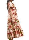 A woman wearing a cream-colored tiered midi dress with a red and green floral print, standing in front of a background of green palm leaves.