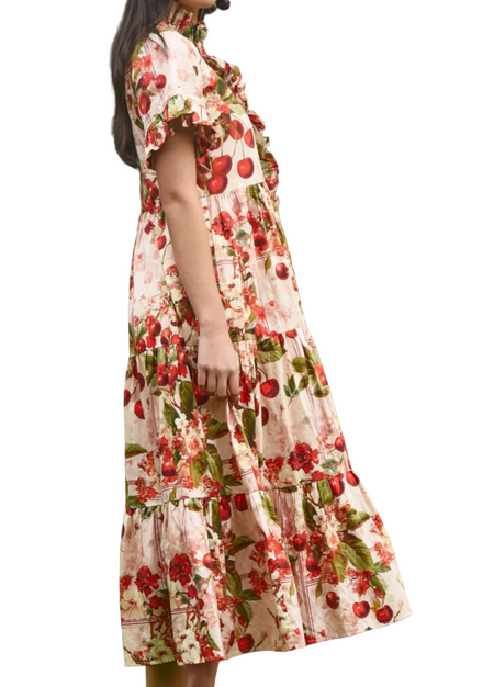 A woman wearing a cream-colored tiered midi dress with a red and green floral print, standing in front of a background of green palm leaves.