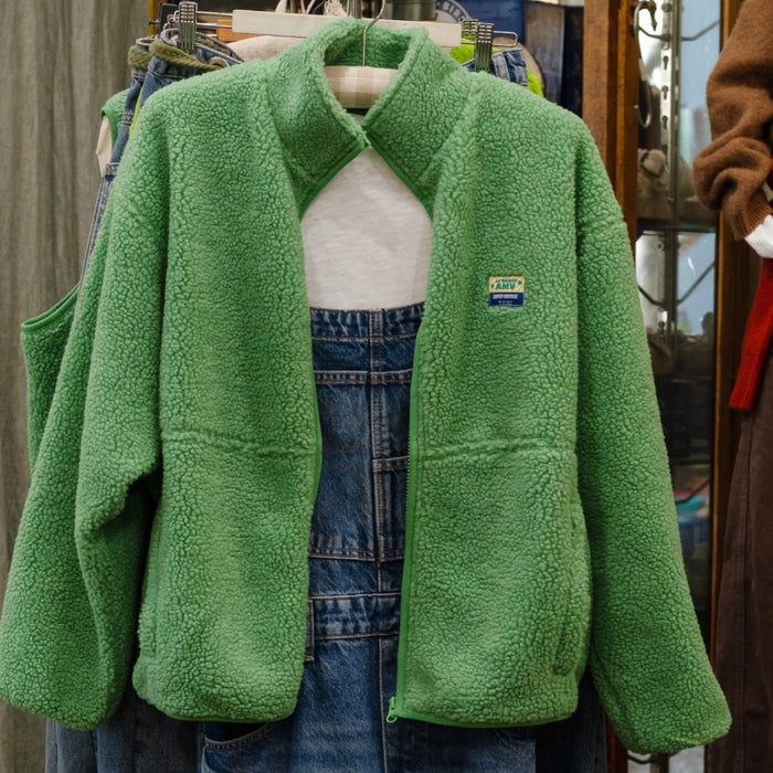 American Vintage green sherpa fleece zip jacket with stand collar, hanging open over denim overalls in the Pomme shop