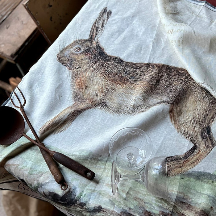 Barbara O'Donovan XL linen tea towel with a hare illustration, styled on a table with a rustic iron ladle, fork and cut-glass coupe