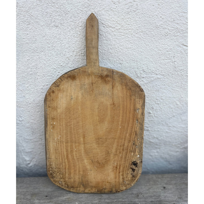 Pale vintage wooden bread board with a pointed handle and worn knife marks, leaning against a whitewashed wall