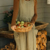 Close-up of the olive gingham linen pinafore dress by Etika, front pocket detail, worn over a knit jumper while holding a basket of apples