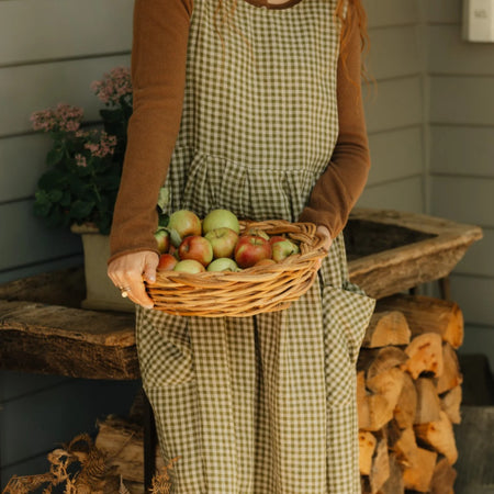 Close-up of the olive gingham linen pinafore dress by Etika, front pocket detail, worn over a knit jumper while holding a basket of apples