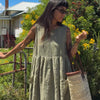 Woman wearing an olive gingham French linen pinafore dress by Etika, styled with a woven straw tote bag