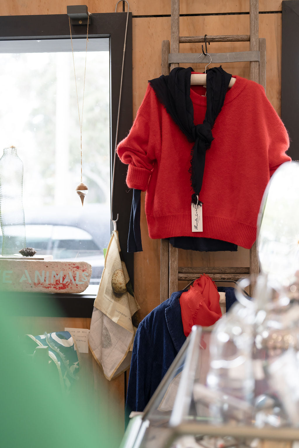 Red Manuelle Guibal knit jumper and black scarf hanging on a wooden ladder by the window inside Pomme at The Studio & Co, Moorooduc